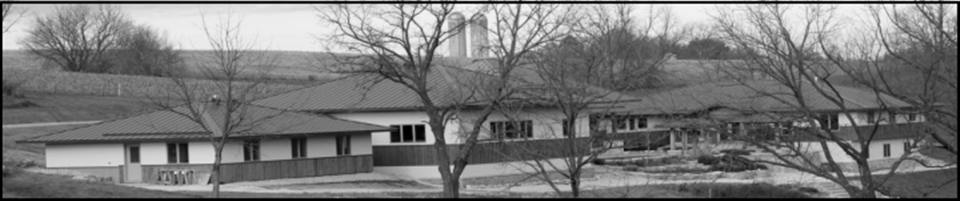 Wide black and white photo of a single-story building with a metal roof surrounded by leafless trees and a stone retaining wall in the background.