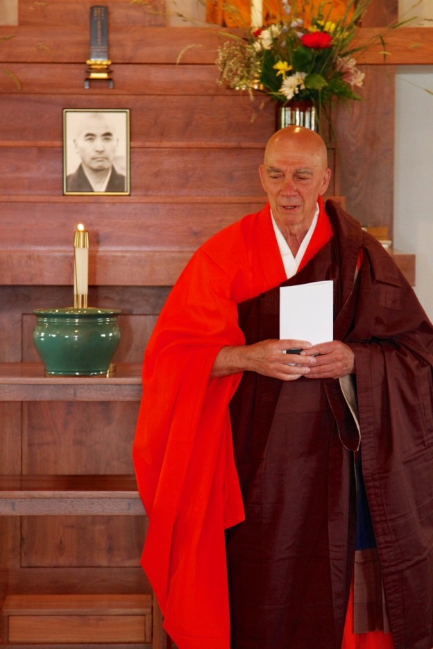 Buddhist monk in orange and brown robes standing in front of a wooden altar with a lit candle, flowers, and a black-and-white portrait.