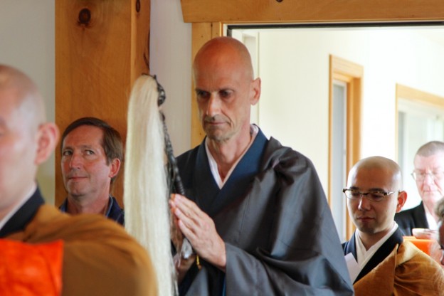 Several men, including one in traditional monk robes holding a large whisk, participating in a ceremonial or religious event indoors.