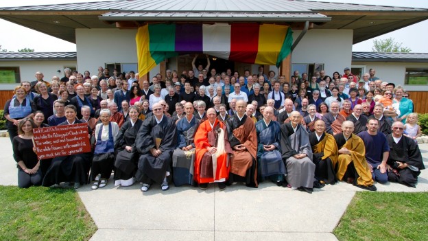 Large group of people including Buddhist monks in robes posing outside a building under a multi-colored banner.
