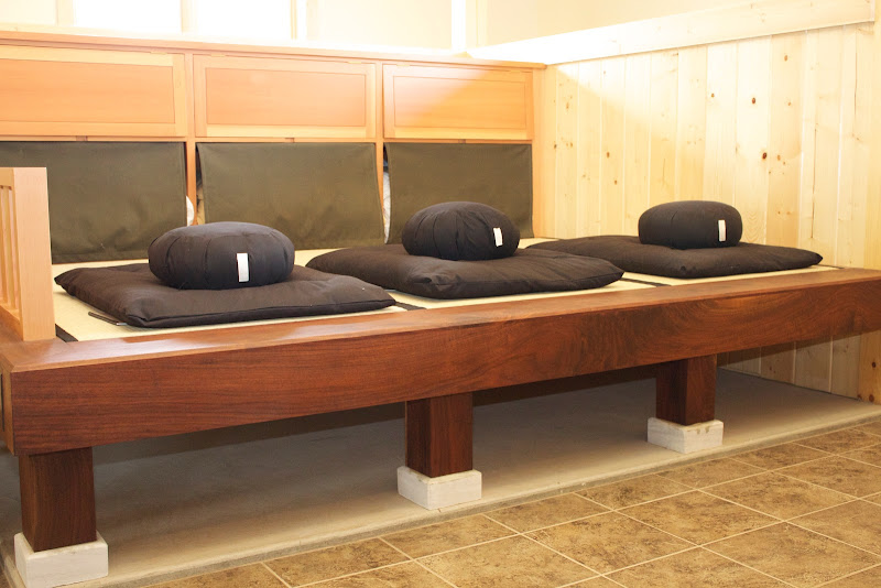 Three meditation cushions on a wooden bench inside a room with wood-paneled walls and tiled floor.