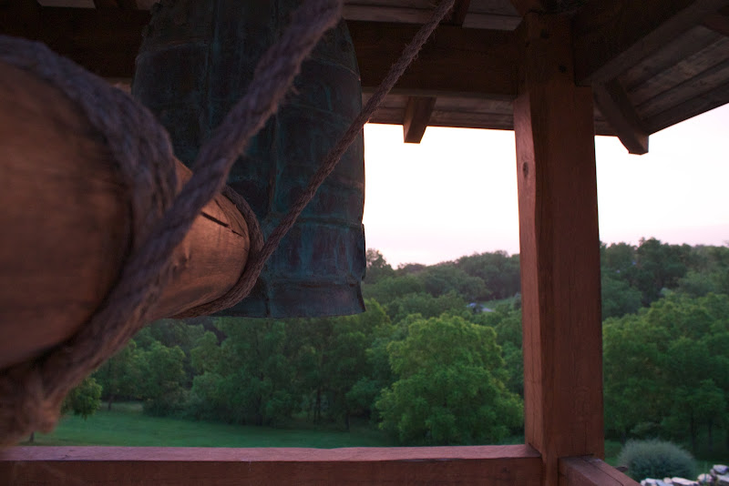 Close-up of a traditional wooden log bell striker suspended by rope next to a large bell, overlooking green trees and a grassy landscape at dusk.
