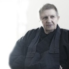 Older man with short gray hair wearing a dark robe-style outfit sitting against a light background.