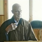 Elderly man wearing glasses and a brown jacket, sitting indoors near a window with a contemplative expression.