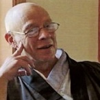 Elderly man wearing glasses and traditional clothing, smiling and resting his cheek on his hand.