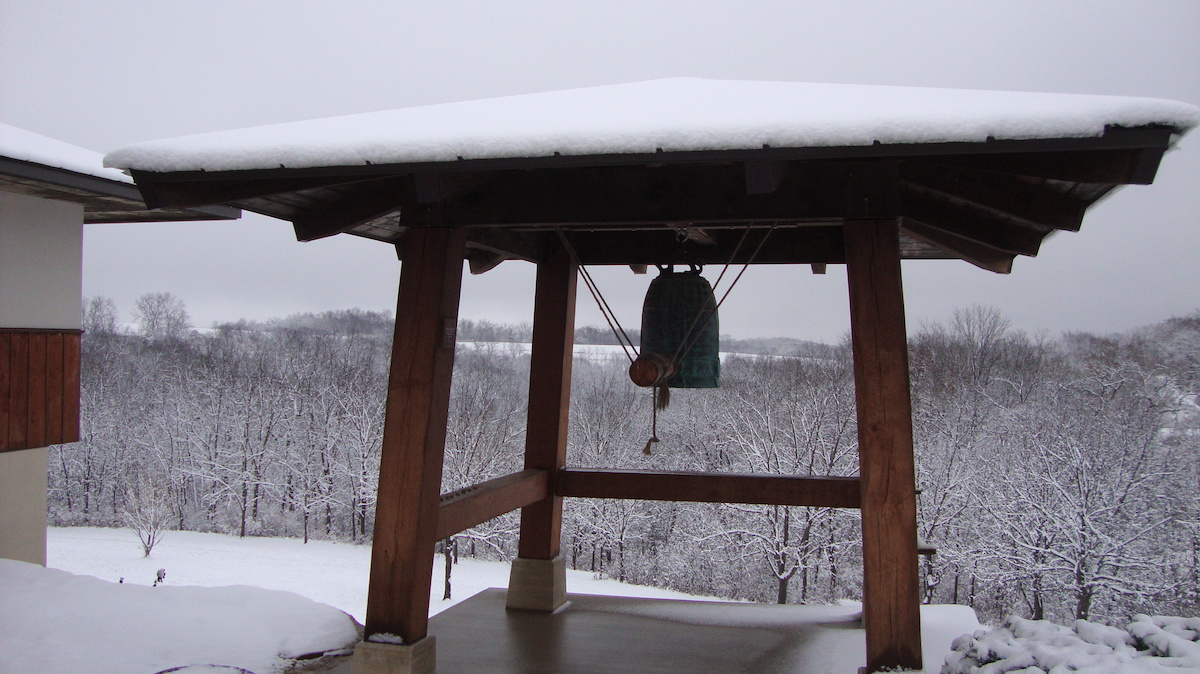 A large bell hanging under a wooden shelter roof covered with snow, overlooking a snowy forest landscape.