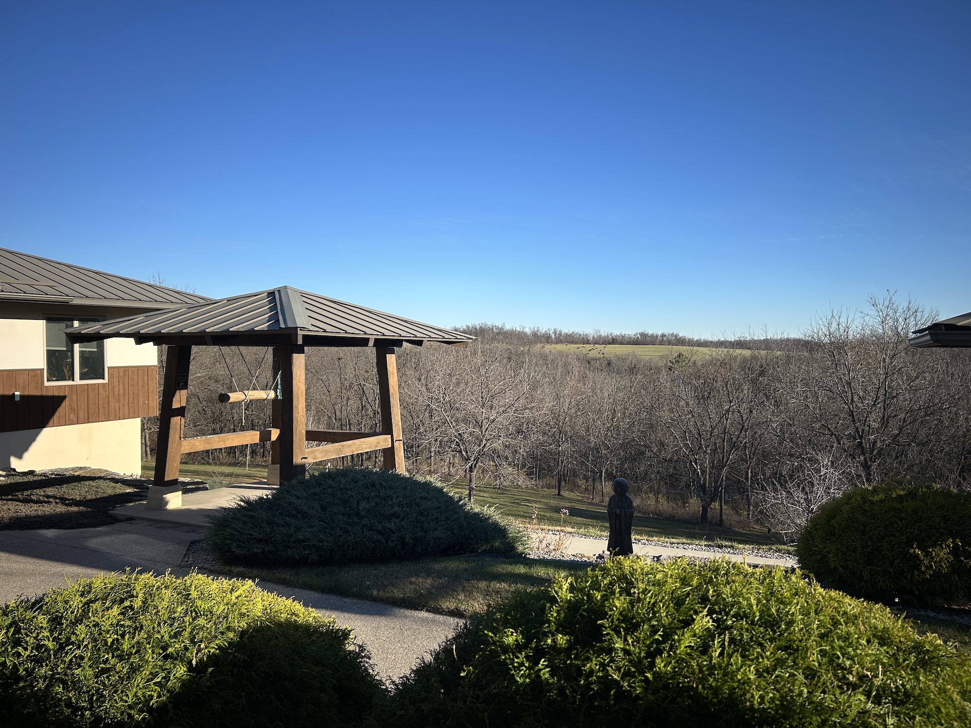 Wooden outdoor bell structure with a metal roof, surrounded by shrubs, with leafless trees and a clear blue sky in the background.