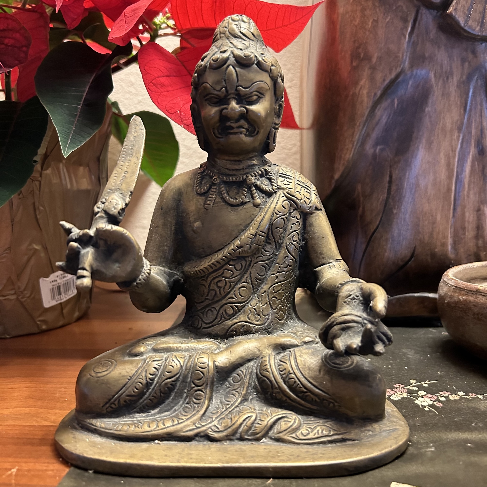 Bronze statue of a seated figure with intricate patterns, holding a small knife in the right hand and a gesture with the left hand, placed on a wooden surface with red poinsettia leaves in the background.
