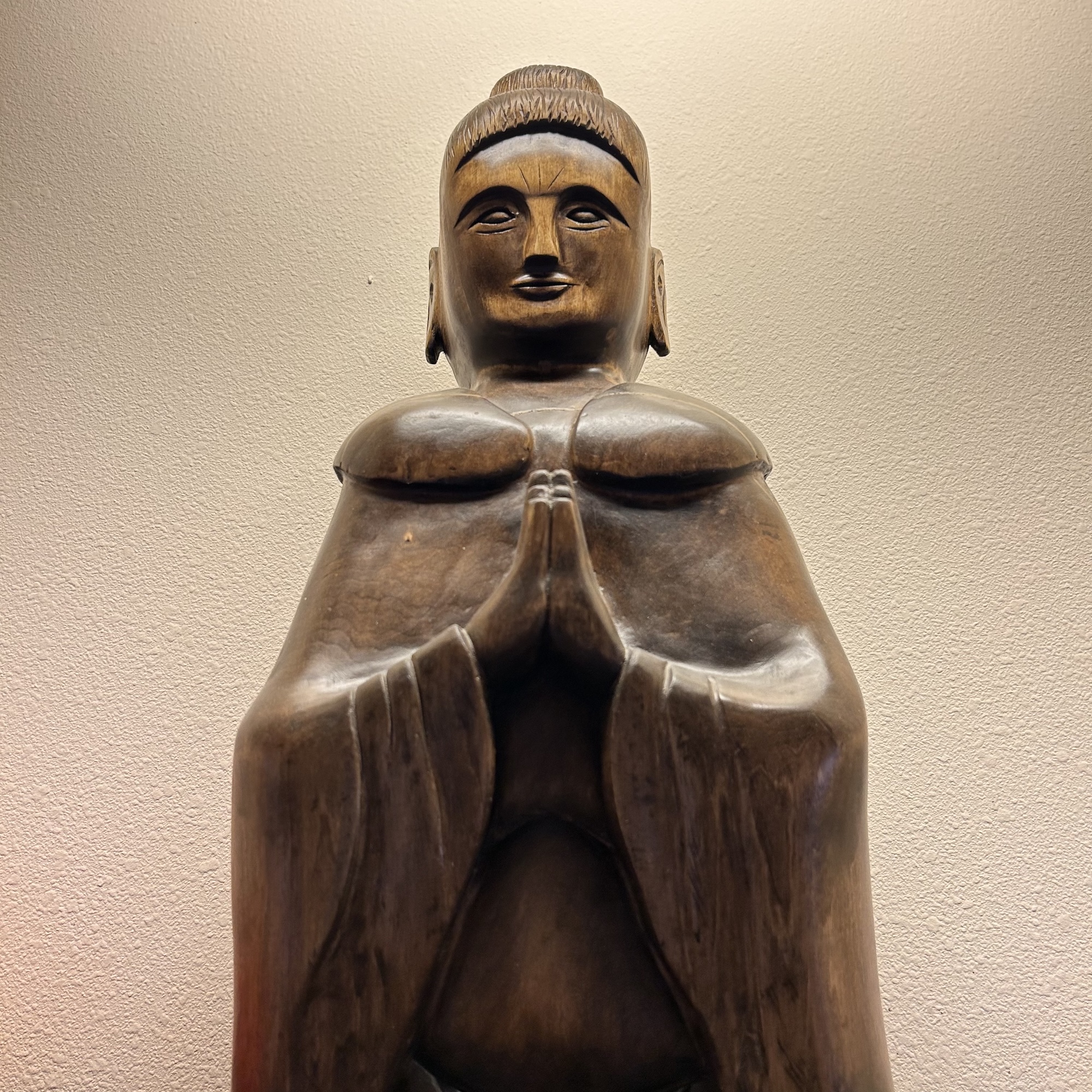 Wooden Buddha statue with hands pressed together in a prayer gesture against a textured light wall.
