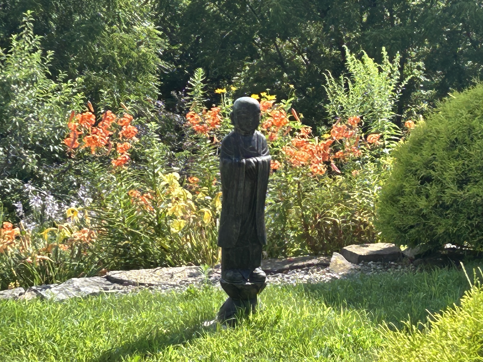 Small dark statue of a standing person in a garden with green grass, orange flowers, and shrubs.