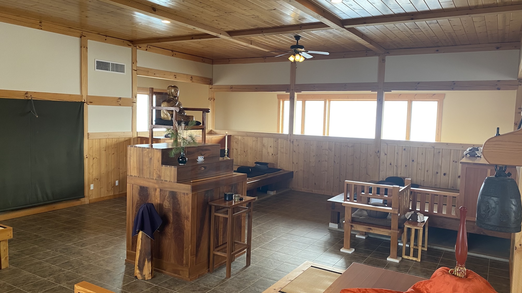 Spacious wooden interior room with a raised platform featuring a seated Buddha statue, low benches with cushions, and a hanging bell.