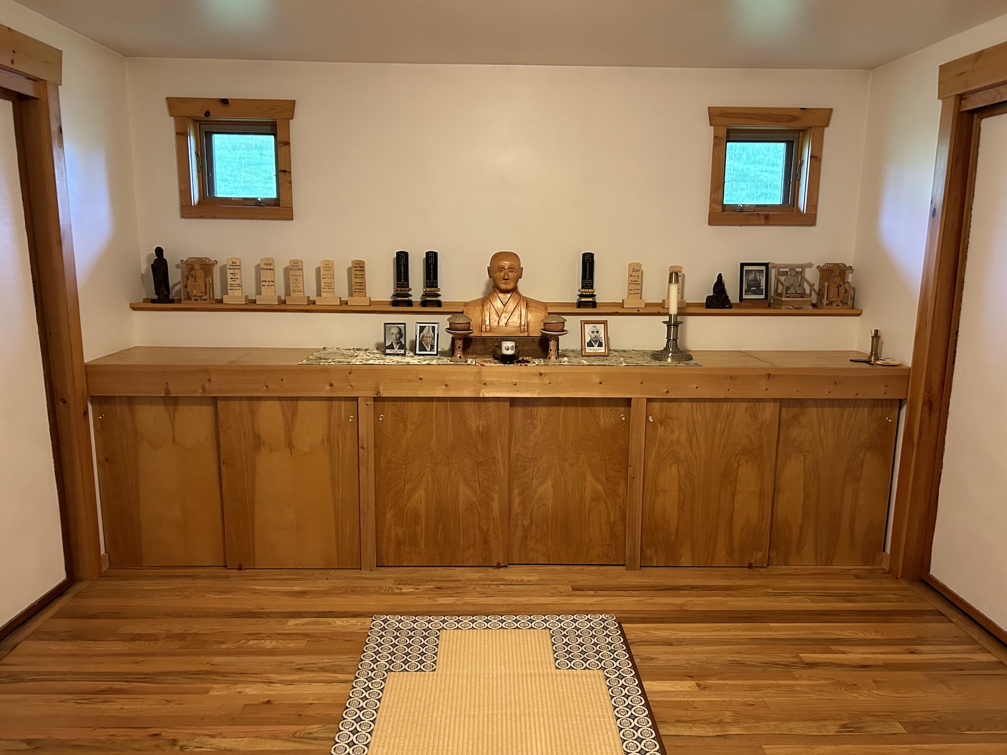 Wooden shrine with bust statue, memorial plaques, framed photos, candles, and small statues on a shelf against a white wall.