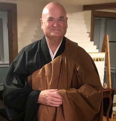 Two people wearing traditional black robes with brown kesa, standing indoors near a wooden door and decorative mirror.