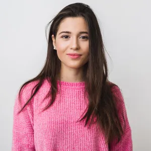 Smiling woman with long dark hair wearing a bright pink knit sweater against a plain white background.