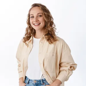 Smiling young woman with curly hair wearing a beige jacket over a white shirt and blue jeans against a white background.