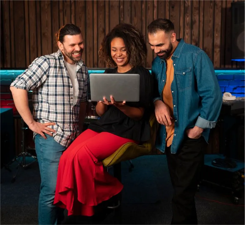 Three musicians smiling and looking at a laptop in a cozy studio with wooden walls and colorful lighting.