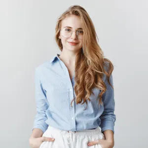 Woman with long wavy blonde hair and glasses wearing a light blue button-up shirt and white pants against a plain background.