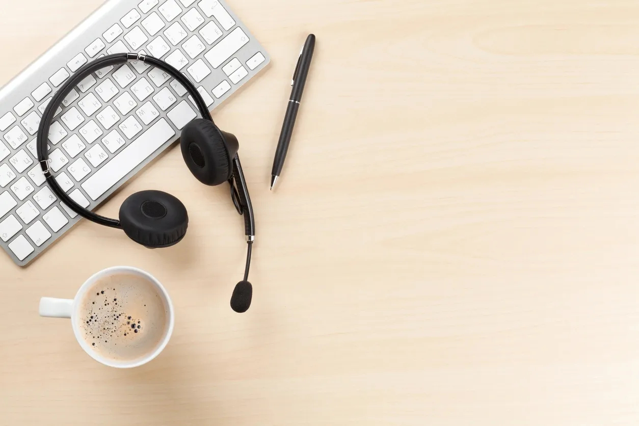 Black headset with microphone, black pen, coffee cup, and computer keyboard on a light wooden desk.