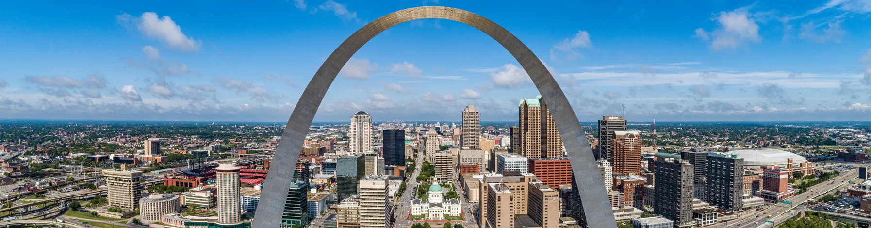 Panoramic daytime view of St. Louis skyline with the Gateway Arch prominently in the center under a partly cloudy blue sky.