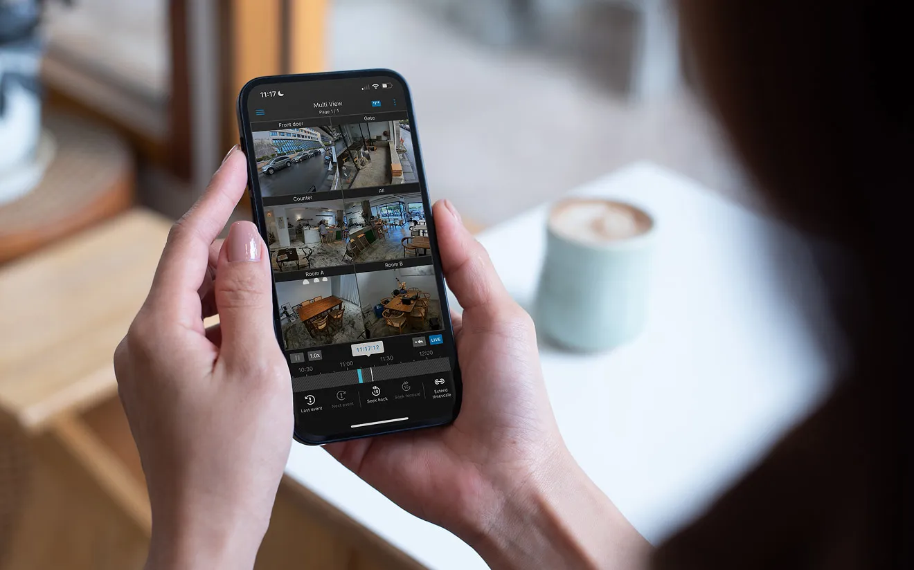 Person holding a smartphone displaying a multi-camera surveillance app showing six different indoor and outdoor views, including front door, gate, counter, and rooms in a cafe or office, with a blurred coffee cup in the background.