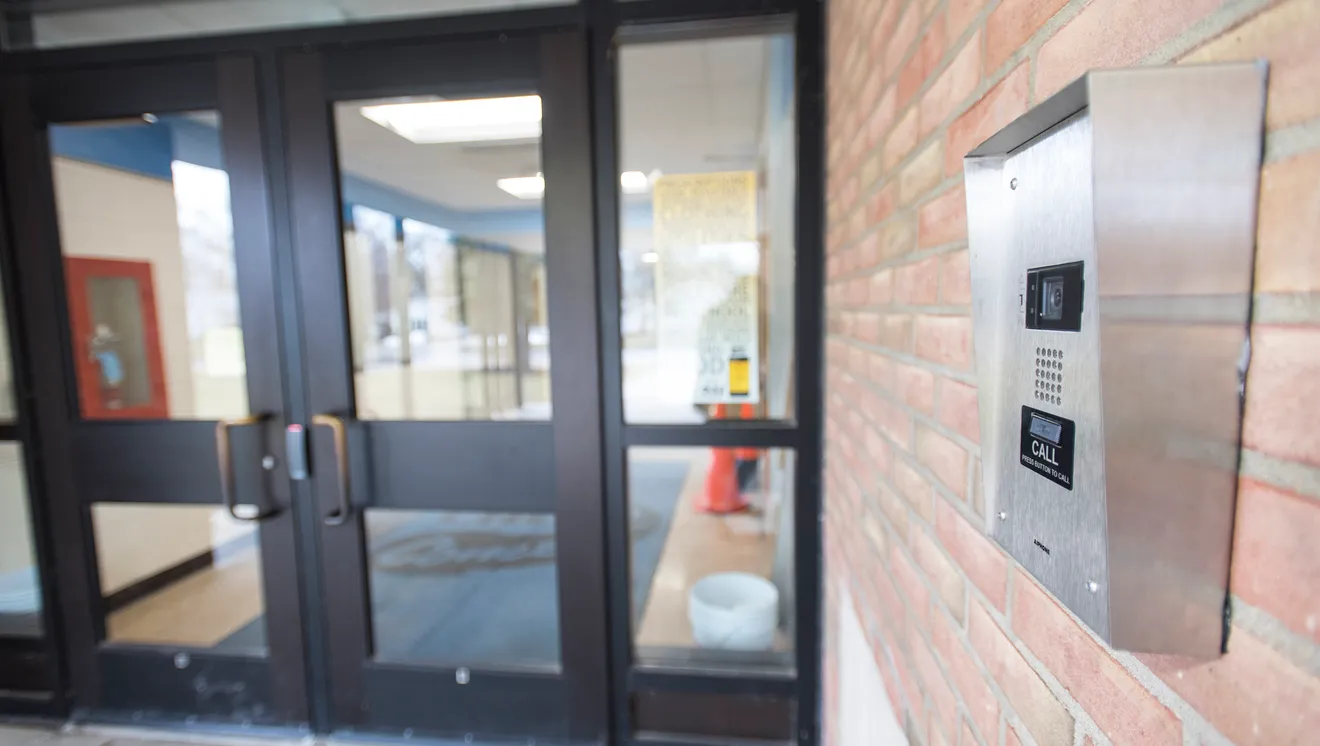 Metal access control intercom panel with a camera, speaker, and call button mounted on a brick wall next to a set of glass double doors.