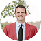 Smiling man with short brown hair wearing a gray suit jacket and reddish tie outdoors with trees in the background.