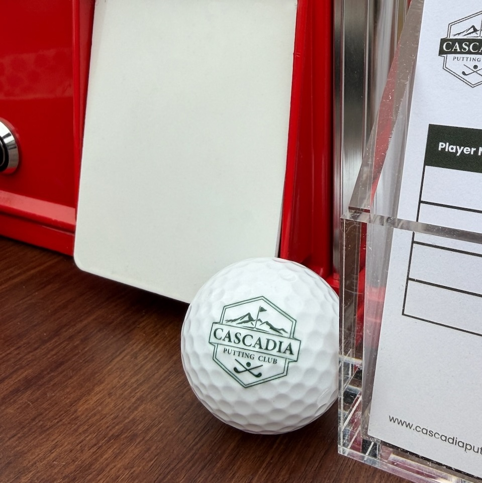 White golf ball with Cascadia Putting Club logo next to a red vintage scoreboard and clear scorecard holder.