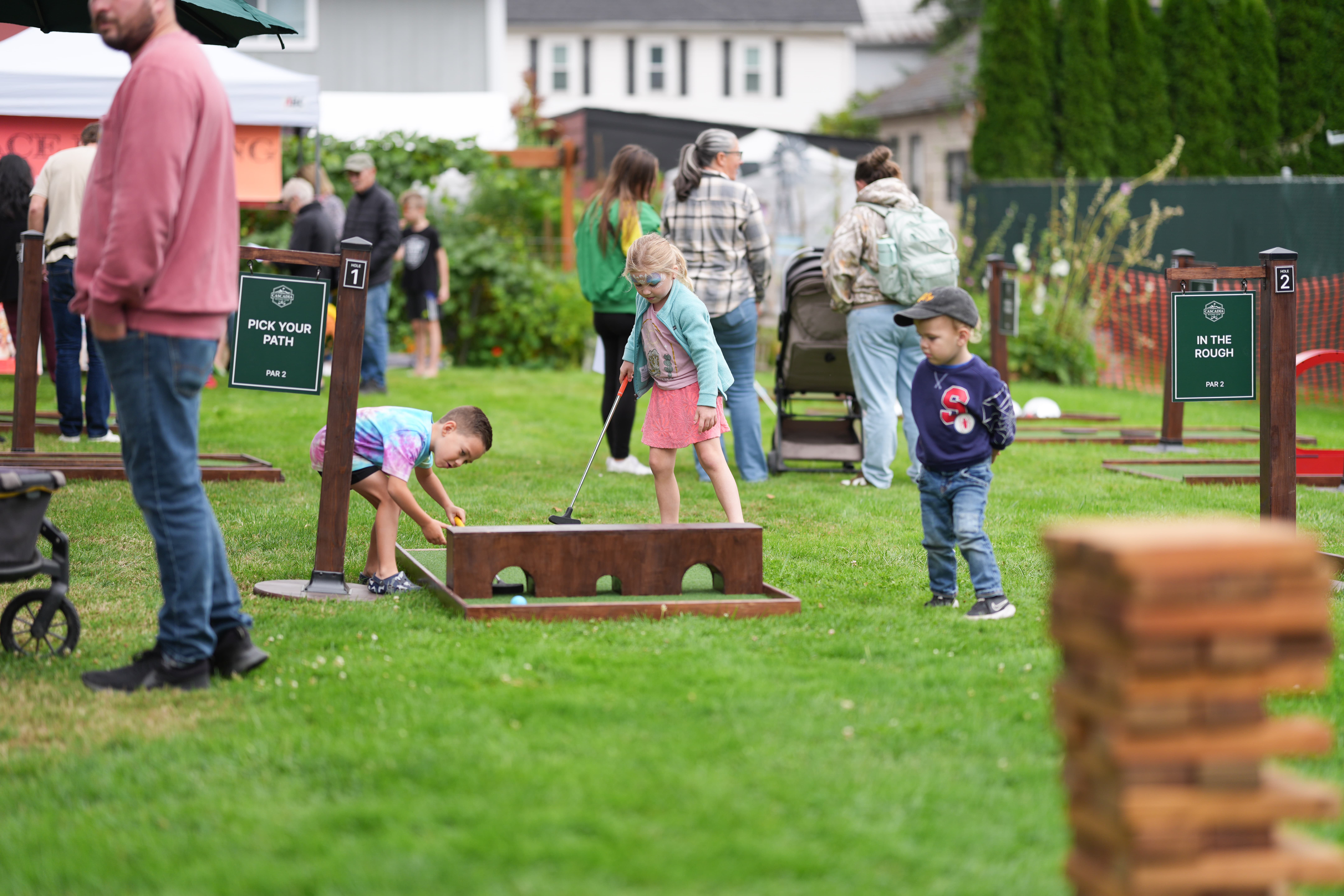 Children playing mini golf outdoors on green grass near signs labeled Hole 1 'Pick Your Path' and Hole 2 'In The Rough' with adults and other kids in the background.