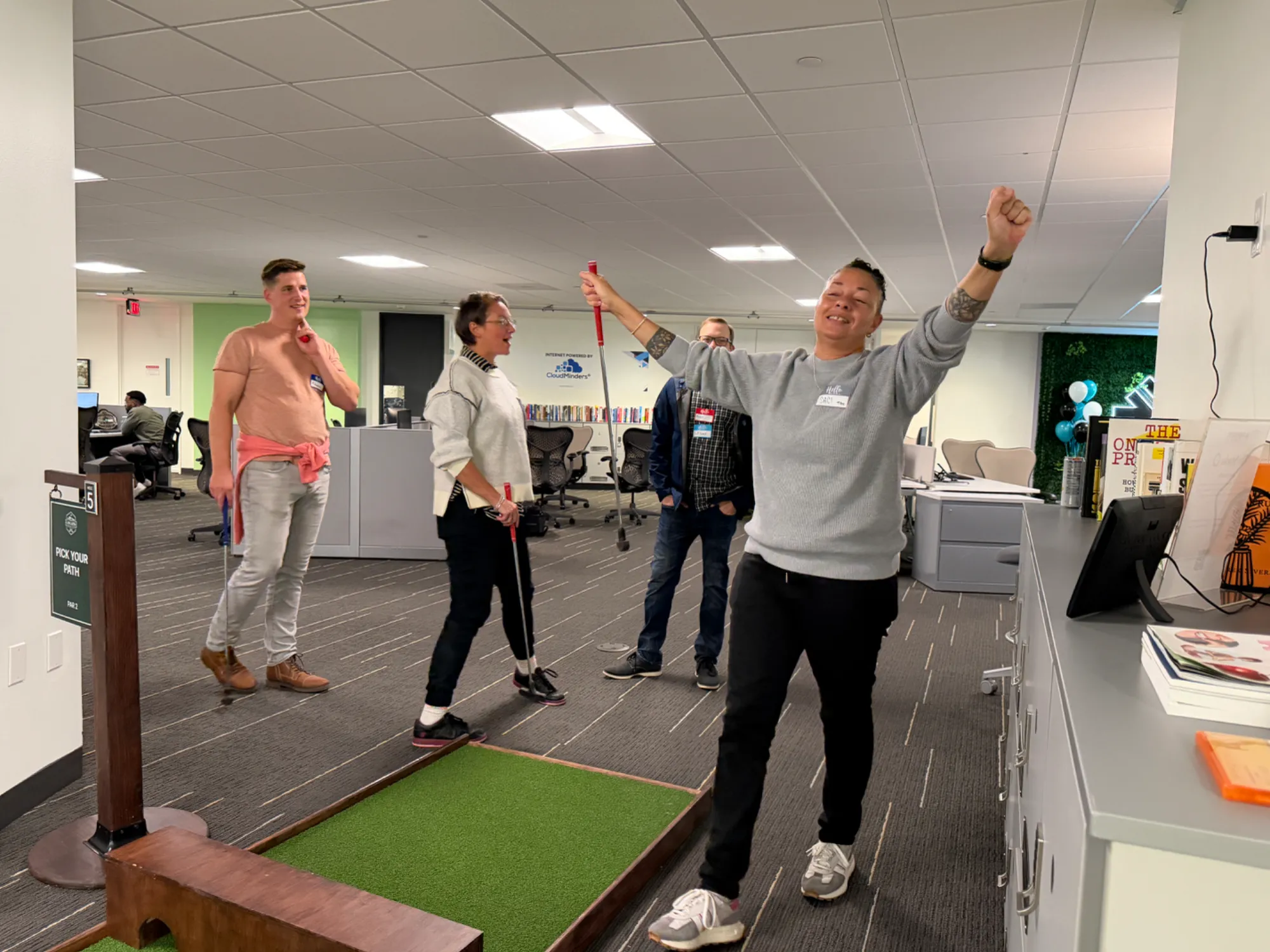Four people playing indoor mini golf in an office space; one person in a gray sweater celebrates with raised arms near the putting green, while three others watch and hold putters.