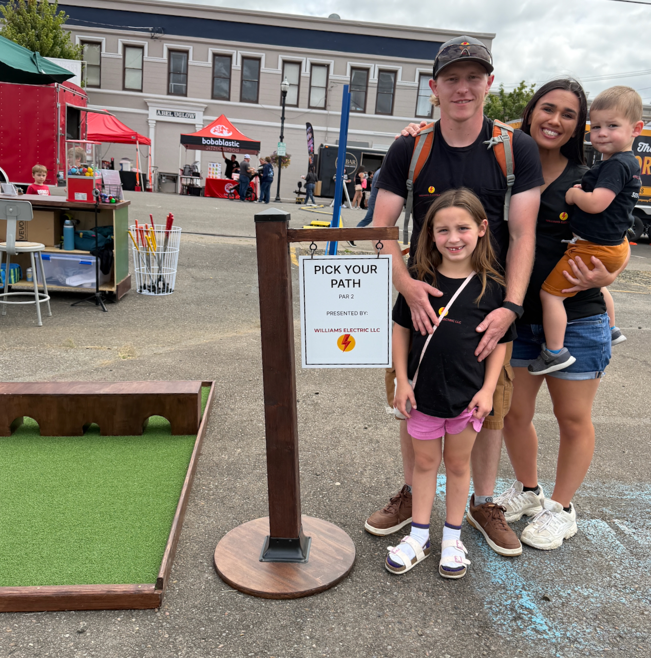 A family of four posing outdoors at an event near a sign that reads 'Pick Your Path Par 2 Presented by Williams Electric LLC'; the family wears matching black shirts with the company logo and stands next to a small mini golf putting green.