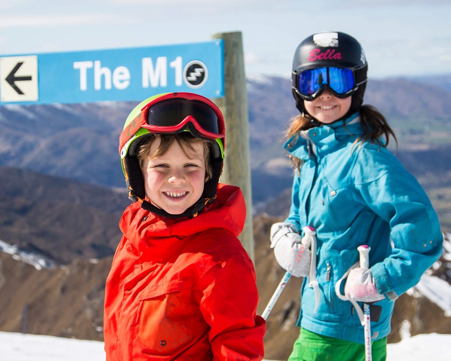Two children standing in front of ski trail sign smiling.