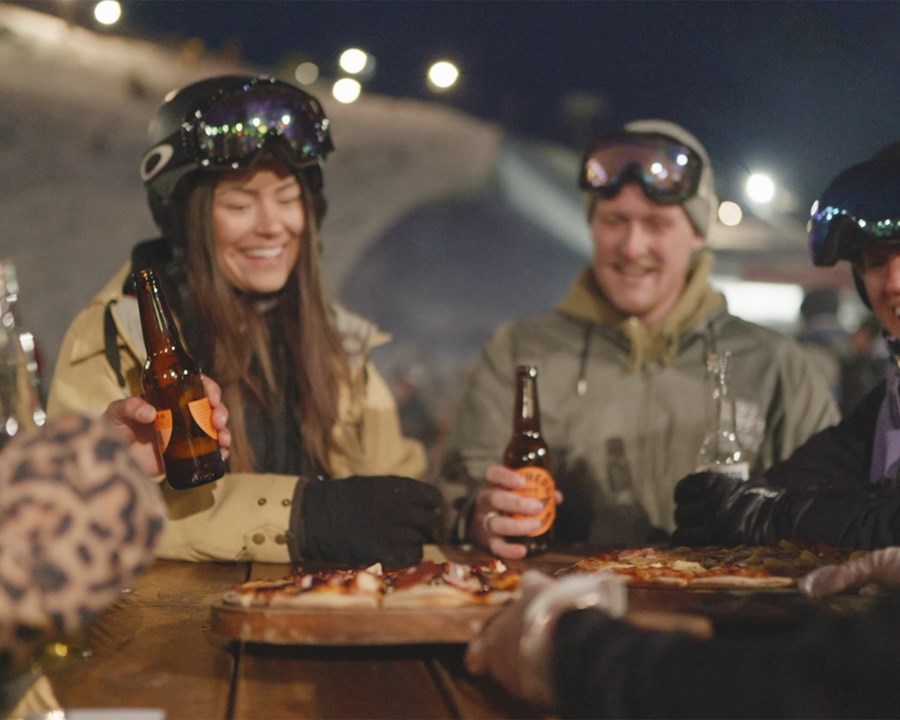 Friends in ski gear, smiling, with drinks in hand and food on the table with a snowcovered slope just behind them.