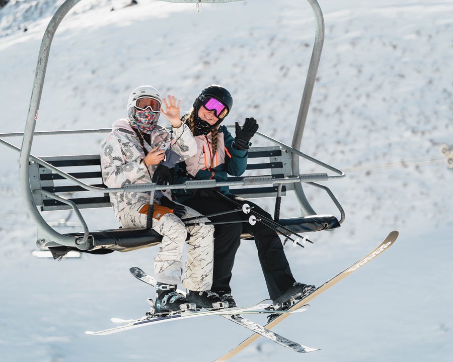 Two women sitting on a chairlift smiling and waving.