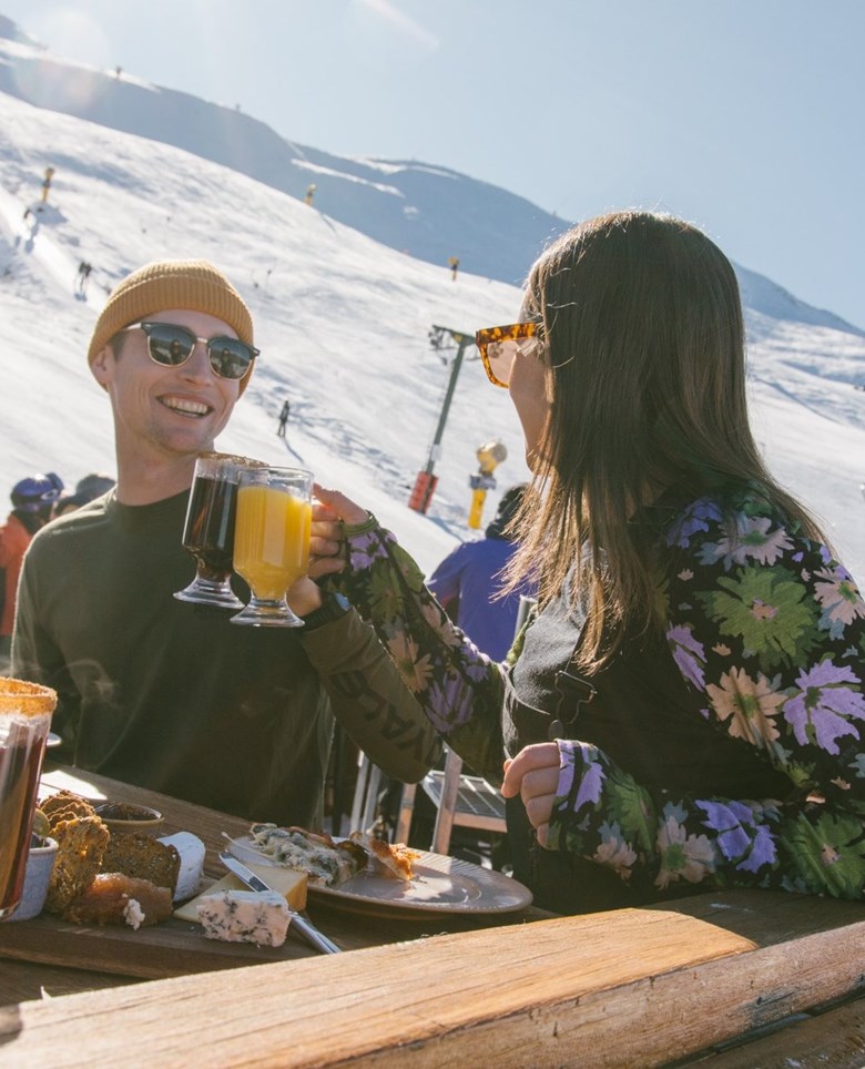 Couple enjoying drinsk & cheeseboard outside in the sun with a snowy mountain slope behind them.