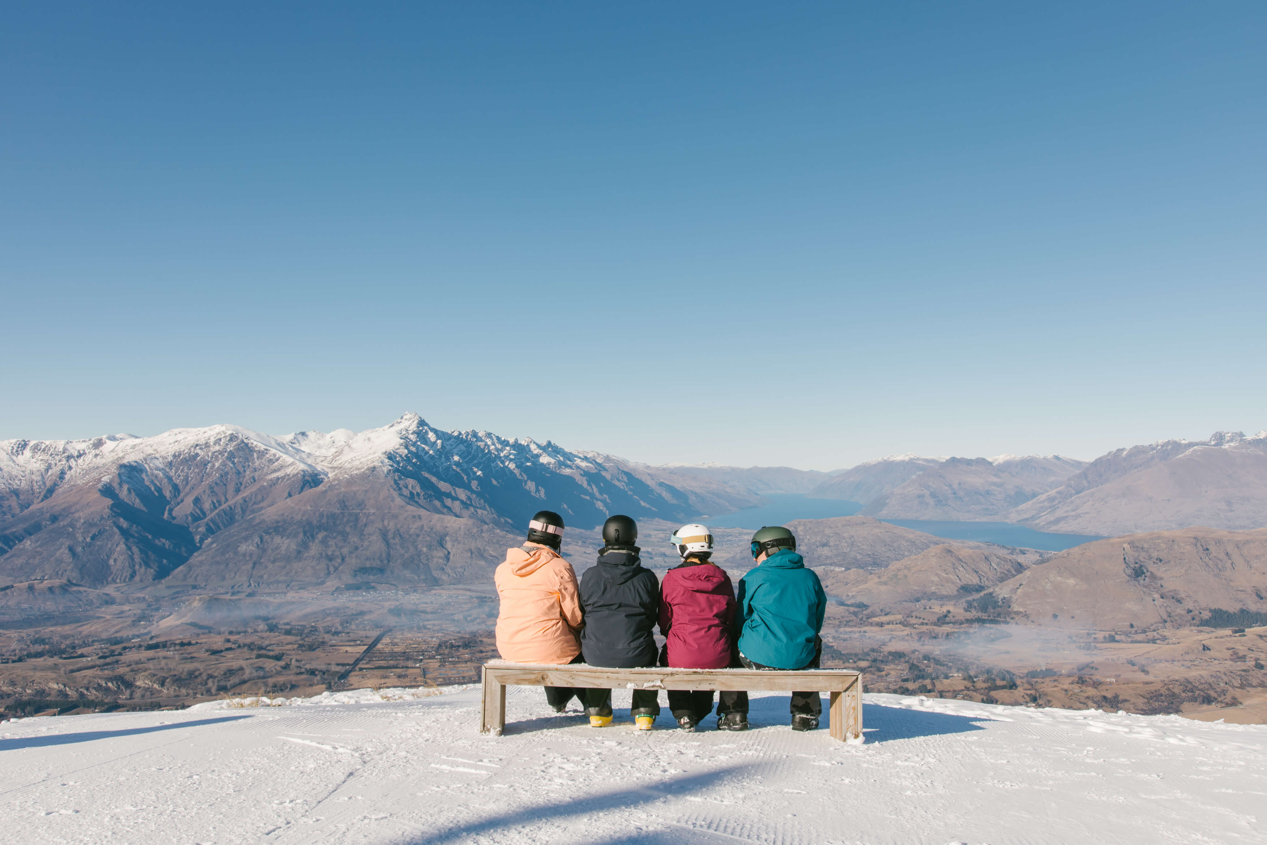 Four people in ski gear sitting on a bench facing snowy mountains and a blue lake under a clear sky.