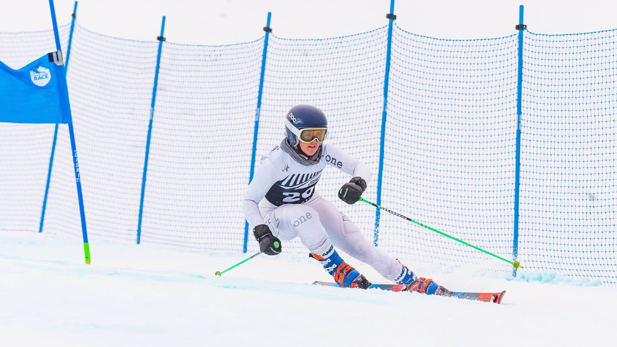 Skier wearing bib 29 racing down a snow slope past a blue race flag and safety net fencing.
