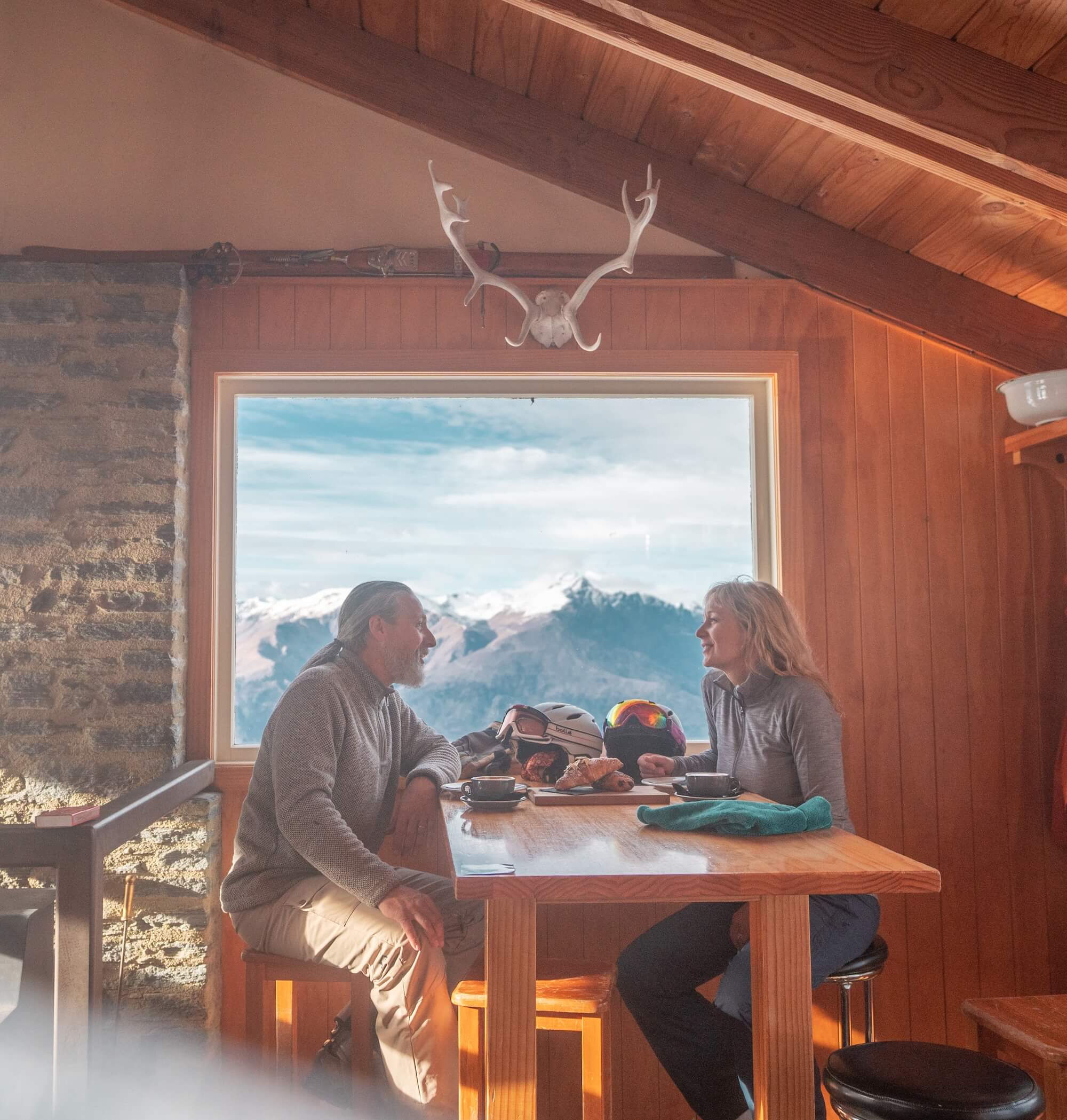 Couple sitting at a wooden table inside a cabin with snowy mountain view through window.