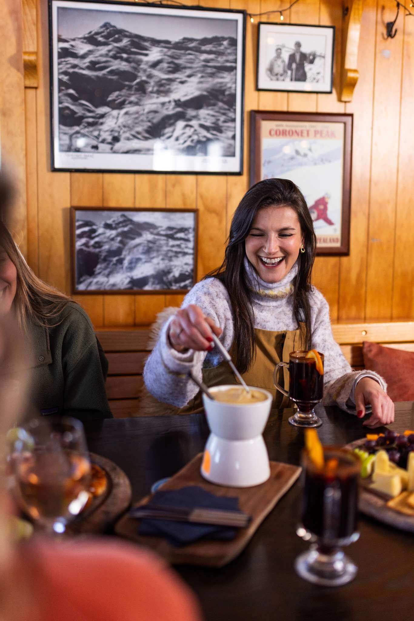Smiling woman stirring a fondue pot at a cozy wooden table with drinks and snacks.