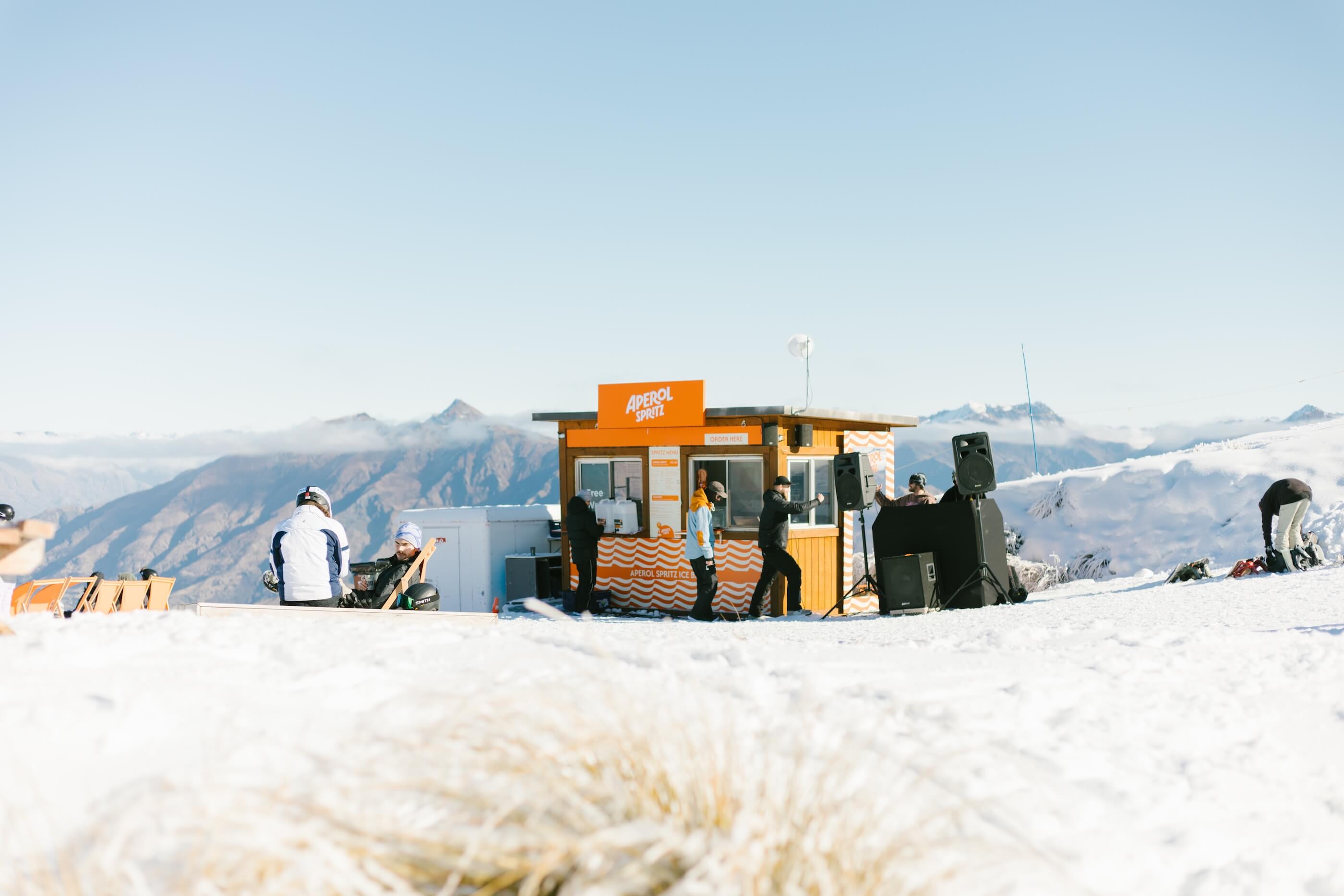 Outdoor Aperol Spritz kiosk on snow with people and mountain views under a clear sky.