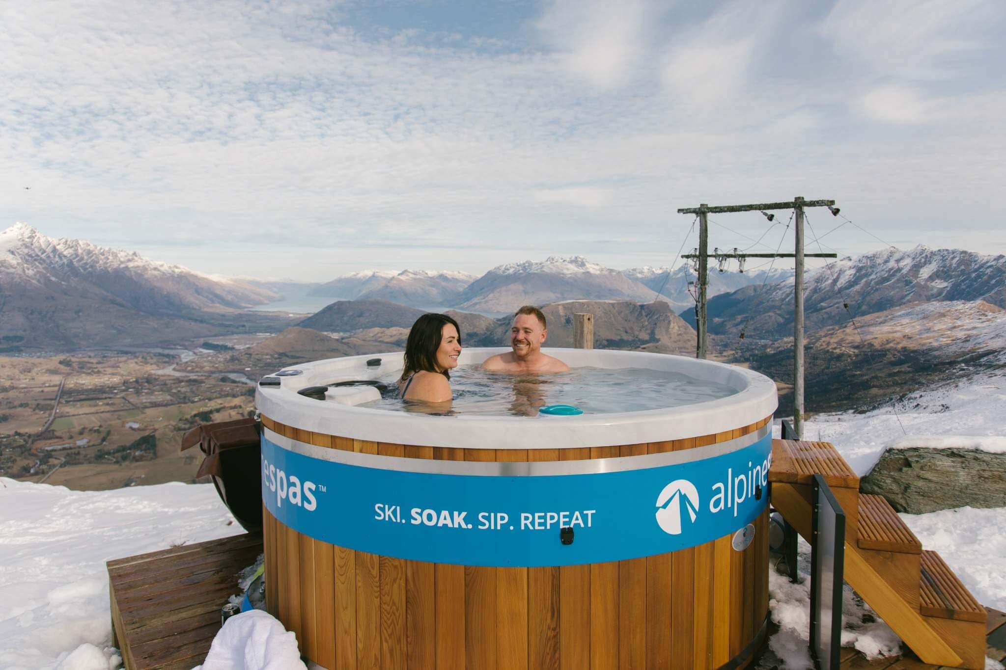 Two people relaxing in a wooden hot tub with snowy mountains and valley in the background.
