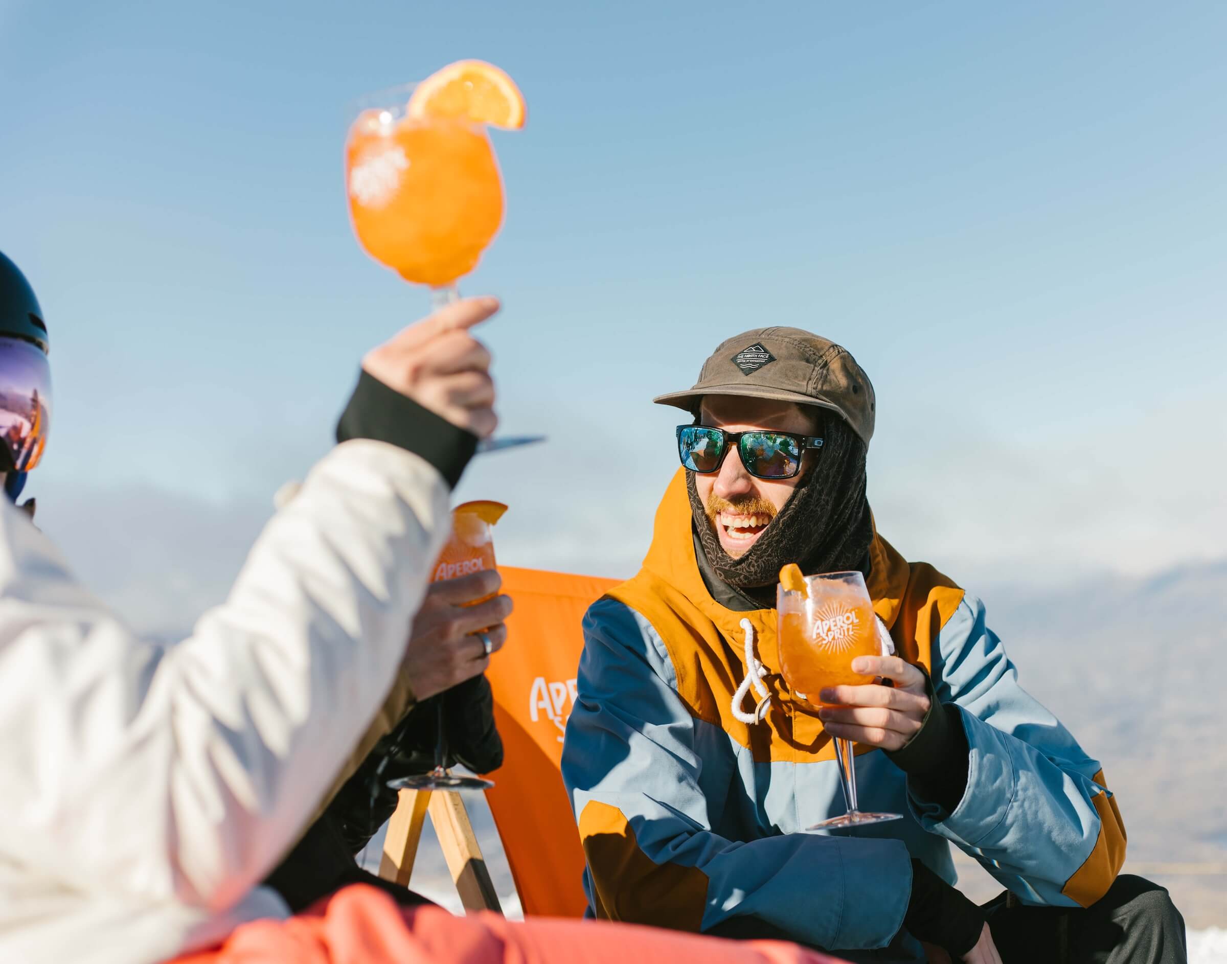 People in winter clothes enjoy Aperol Spritz drinks outdoors with snowy mountains in the background.