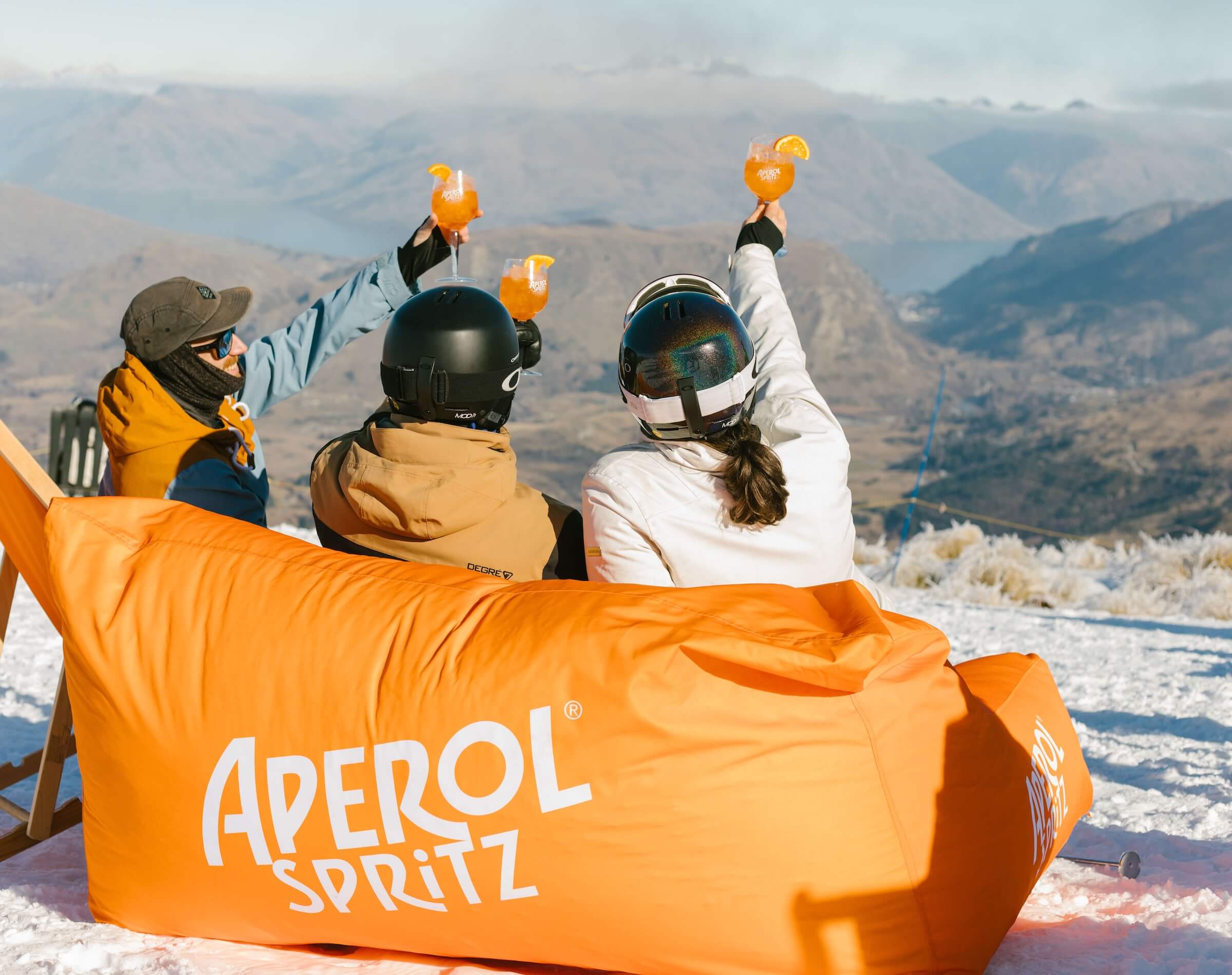 Three people in winter gear toasting Aperol Spritz drinks outdoors with mountain views and snow.