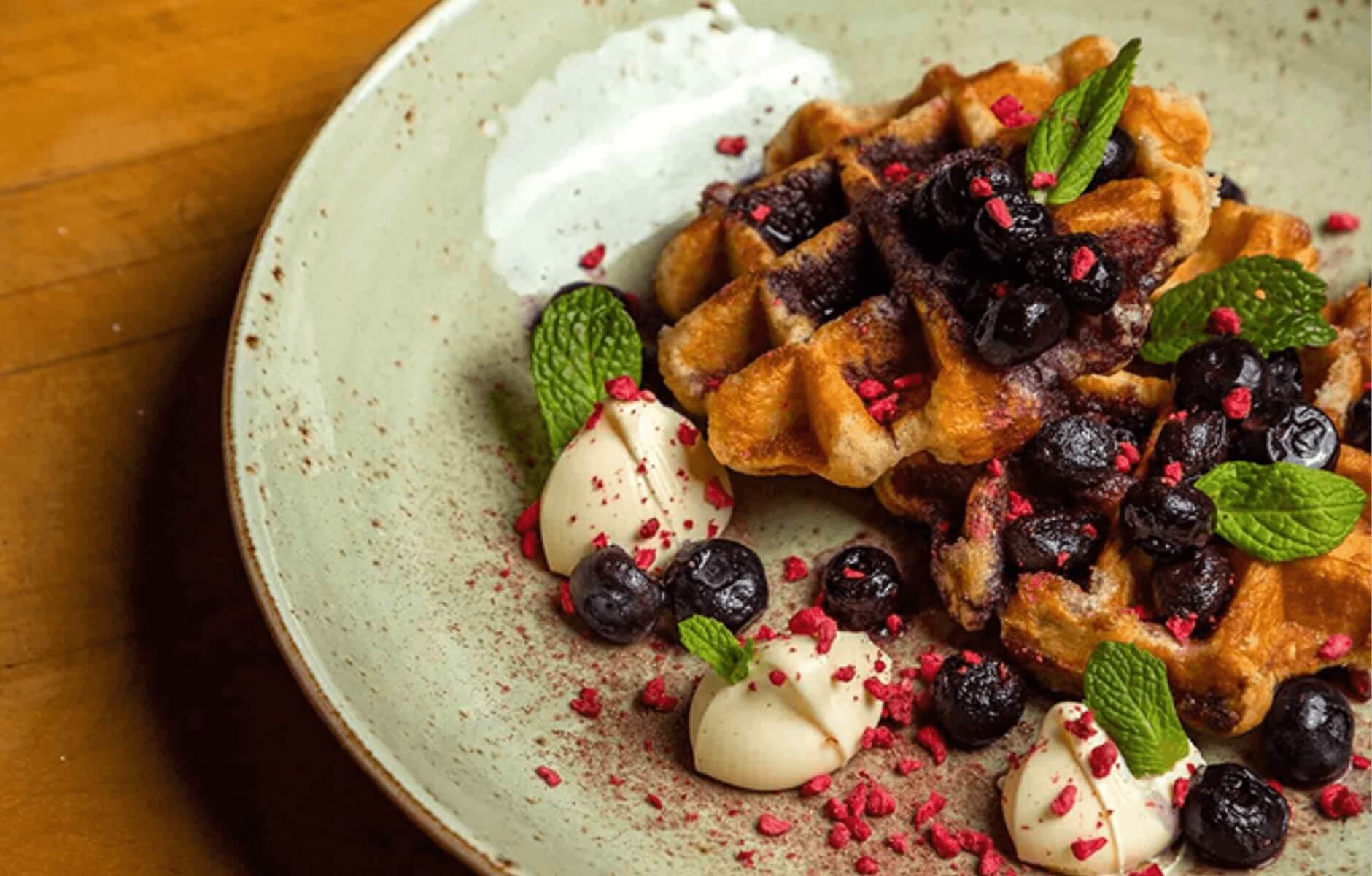 Plate of golden waffles topped with blueberries, mint leaves, cream dollops, and red berry crumbs.