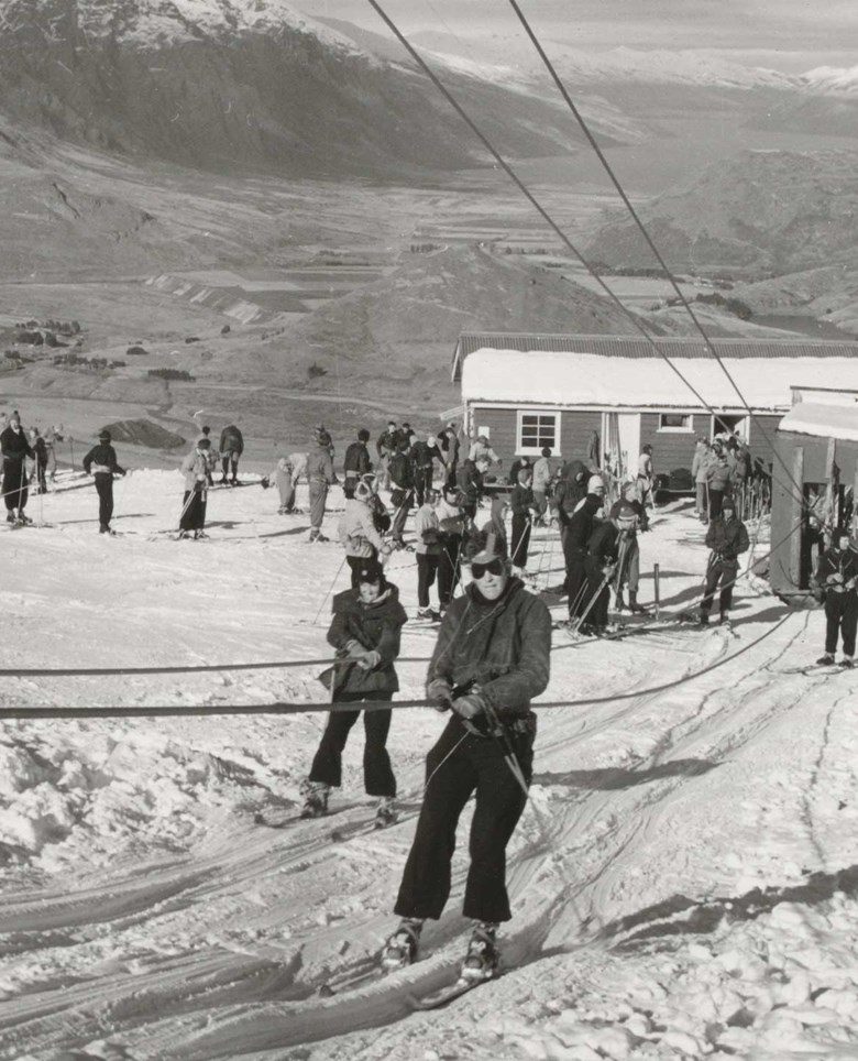 Vintage black and white photo of skiers using a ski tow at a snowy mountain resort with a lodge.