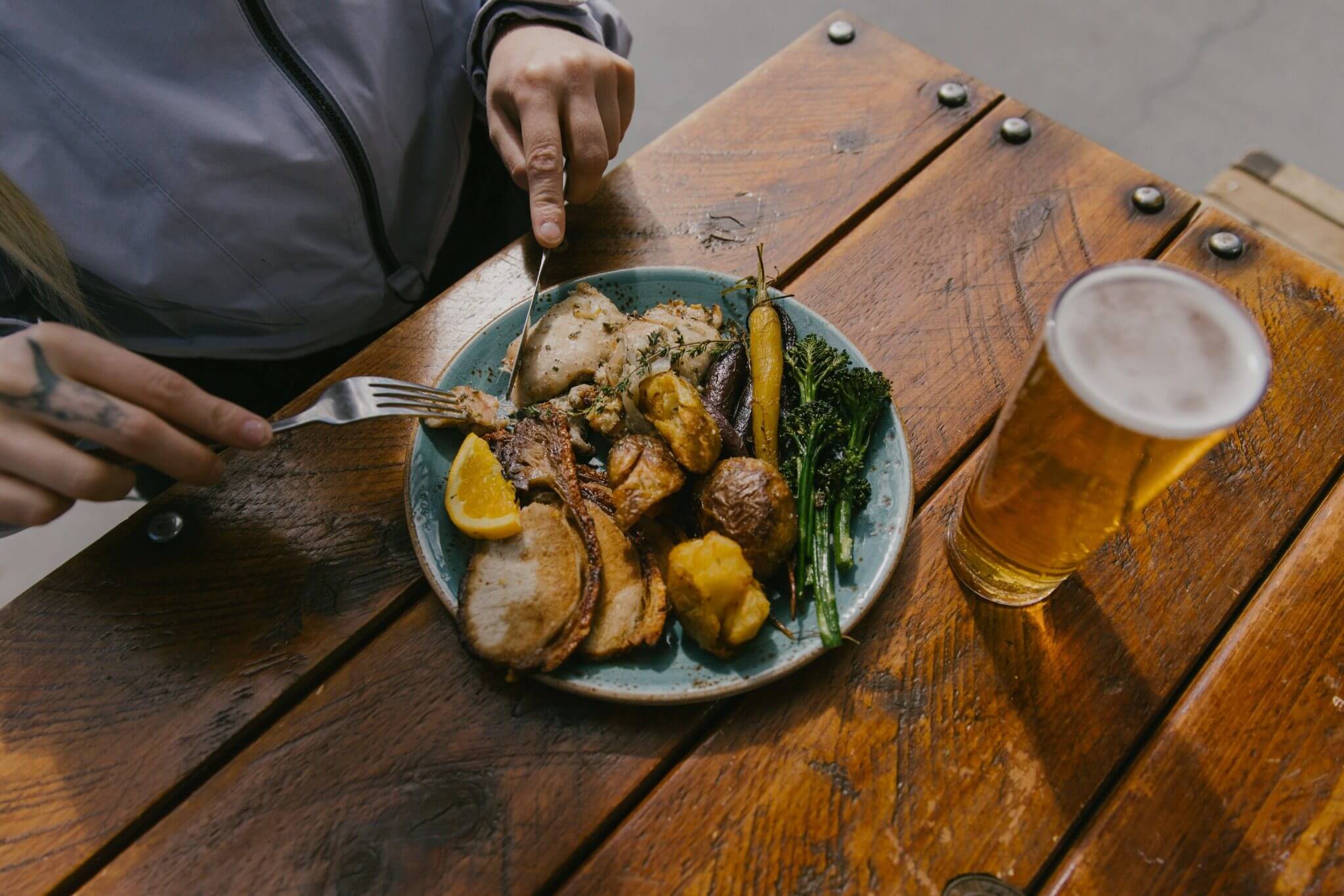Person cutting meat on a plate with roasted vegetables, lemon wedge, and a glass of beer on wood table.