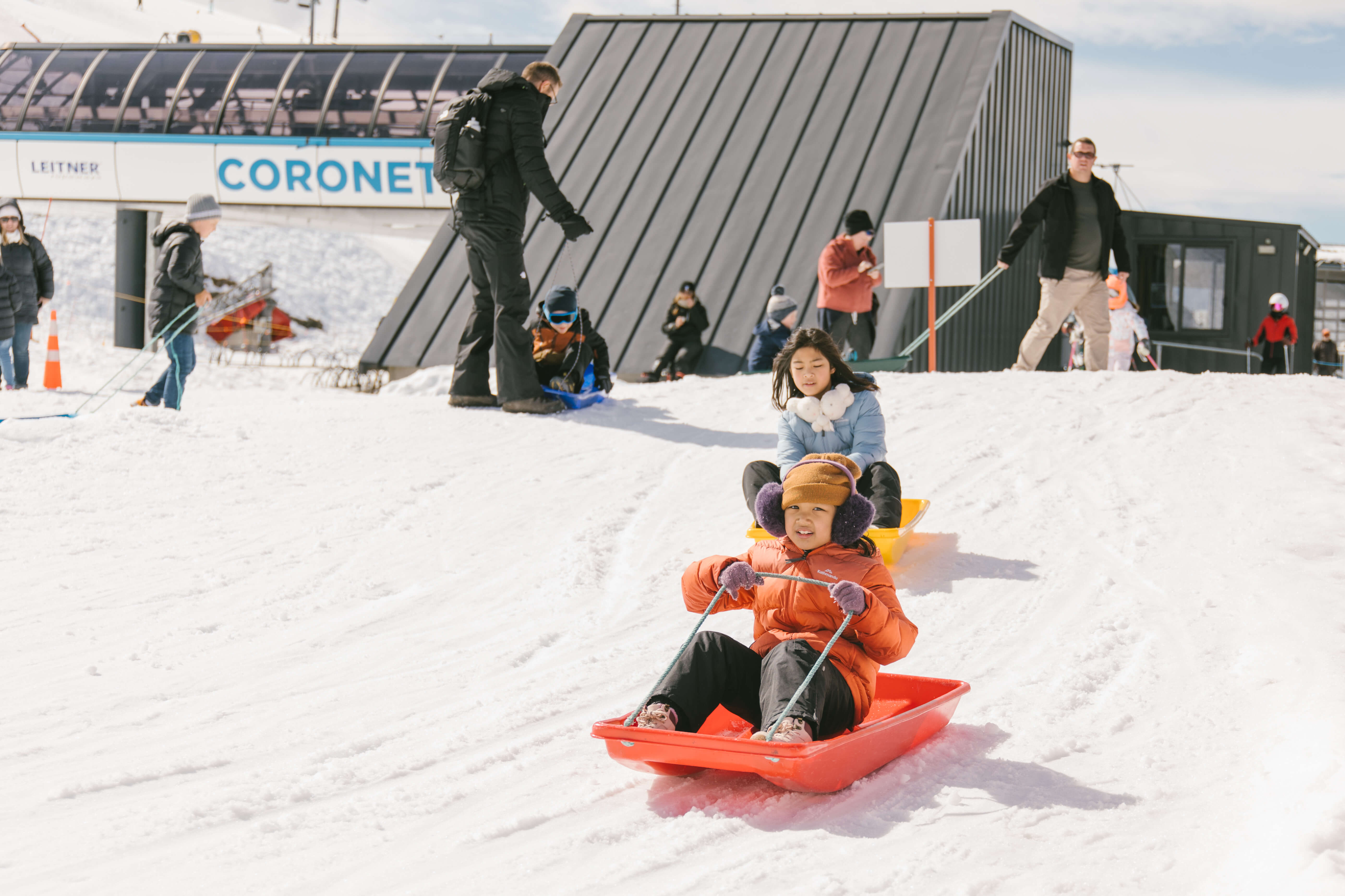 Two children sledding down a snowy hill on red and yellow sleds at a ski resort.