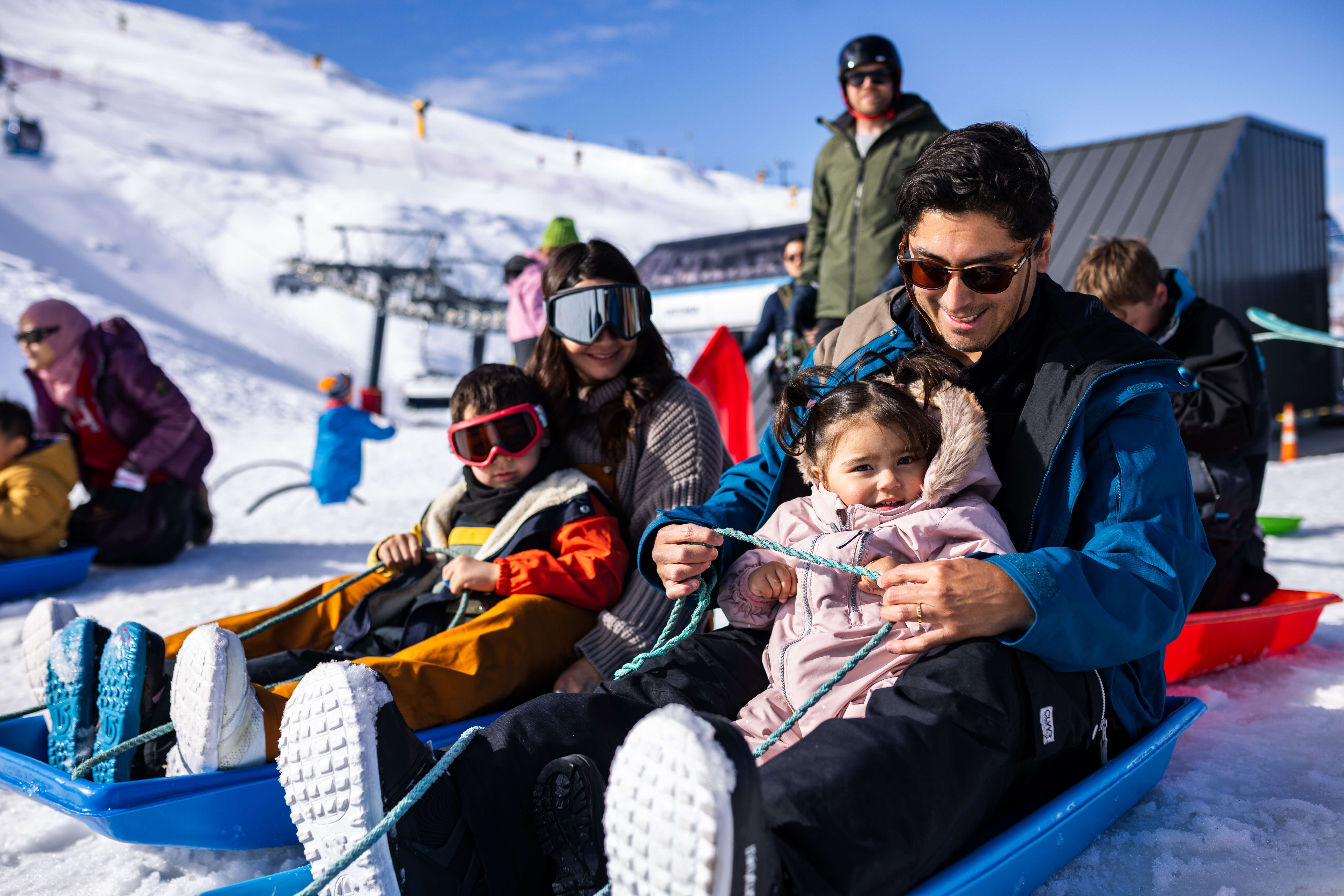 Family with kids wearing winter clothes sledding on snow with ski lifts and slopes in background.