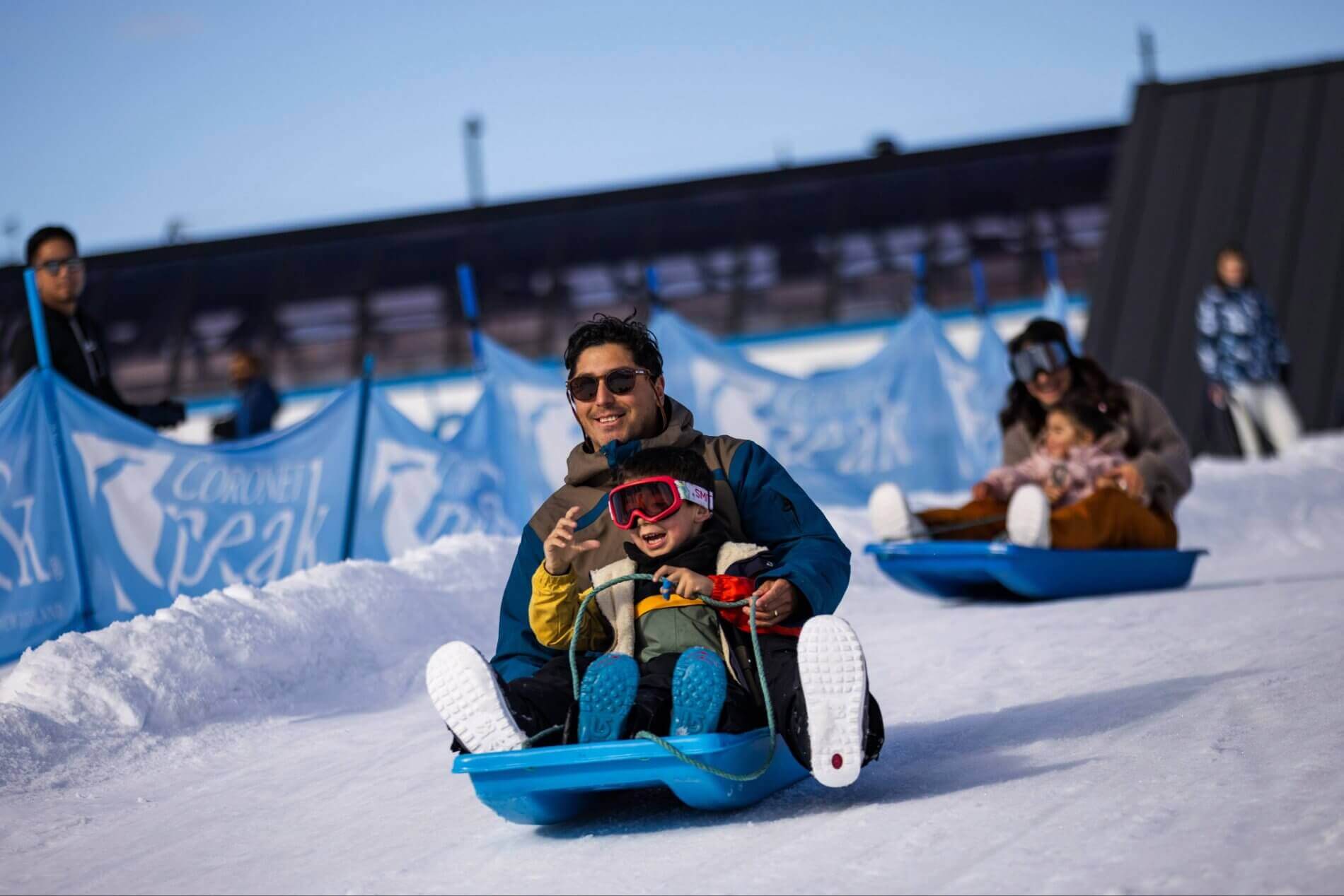 A man and child sledding down a snowy hill on a blue sled, both smiling and wearing winter gear.