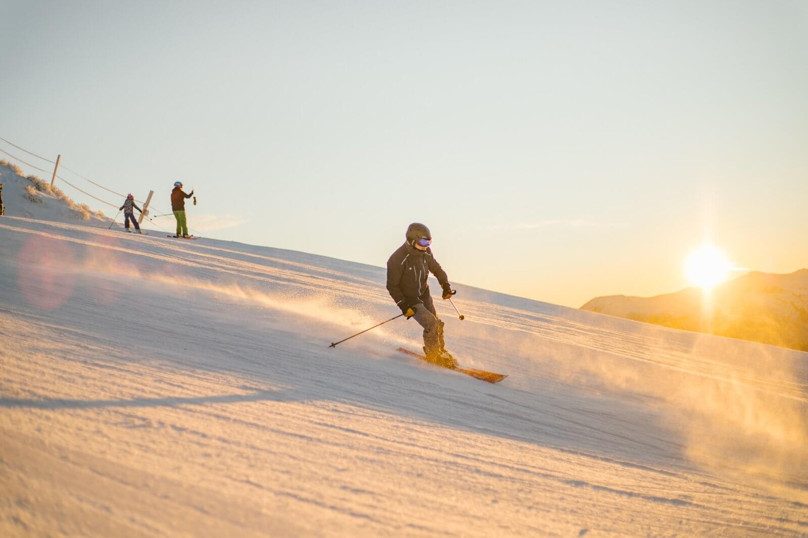 Skier carving fresh snow on a slope at sunrise with two others standing in the background.