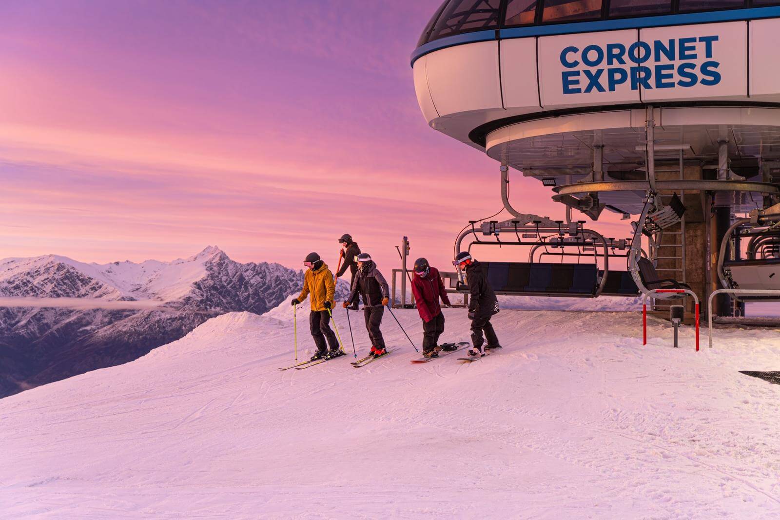 Five skiers and snowboarders at Coronet Express lift with snowy mountains and sunrise sky.